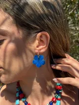 Aro de acero quirúrgico con dije en forma de flor pintado a mano en color azul con acabado brillante. Consta de una pequeña argolla dorada de la que pende un medallón floral artesanal.