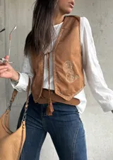 Chaleco de gamuza color camel con bordados decorativos en hilo claro y cierre frontal mediante lazos con borlas. Presenta un diseño de inspiración boho con terminaciones delicadas.