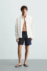 Bañador largo negro con cintura elástica ajustable mediante cordón. Presenta un panel lateral con estampado de cuadros en tonos azul y negro. Incluye bolsillos frontales y un bolsillo trasero con solapa.