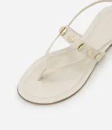 Sandalia plana color beige con tiras finas y tira central decorada con colgantes metálicos dorados en forma de conchas marinas. Cuenta con cierre de hebilla ajustable en el tobillo.