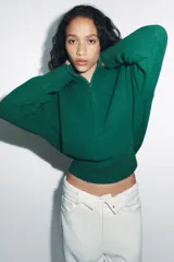 Jersey de punto de color beige con cuello subido y cierre frontal con cremallera metálica. Presenta mangas largas con puños acanalados y diseño de corte holgado.