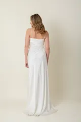 Vestido largo blanco de satén de seda natural, con breteles finos, escote recto, drapeado en el busto, tajo lateral y cola corta. Presenta aplique de flor de organza bordada en un costado.