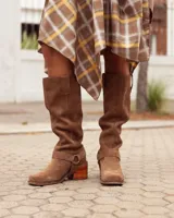 Botas de caña alta confeccionadas en cuero gamuzado color marrón. Presentan un diseño de estilo western con puntera cuadrada, taco de madera y una tira decorativa con estribo metálico desmontable en la zona del tobillo.