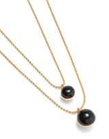 Collar dorado de doble cadena con dije de cristal negro. La cadena superior es más corta y tiene un dije de cristal pequeño, mientras que la cadena inferior es más larga y tiene un dije de cristal más grande.