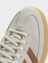 Zapatillas Adidas Handball Spezial de gamuza en tono gris claro, con las icónicas tres tiras laterales y refuerzo en el talón con textura de piel de serpiente en color marrón. Presenta suela de goma color caramelo y detalles dorados en el logo lateral y la lengüeta.