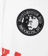 Hoodie blanco con capucha y cordones ajustables, confeccionado en algodón con felpa interior. Presenta estampado frontal con la palabra 'BROOKLYN' en color rojo, un parche circular con la imagen de Notorious B.I.G. y una firma bordada. Cuenta con bolsillo canguro y puños ajustados.