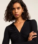 Body negro de rib, con cuello polo, escote en V, mangas largas y frunces en el busto. Cierre inferior con botones a presión.