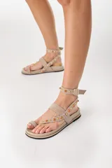 Sandalias planas de cuero marrón, con diseño de tira al dedo (toe-post), tira horizontal en el empeine y tira ajustable en el tobillo. Tienen apliques de tachas metálicas doradas y piedras naranjas a lo largo de las correas.