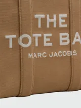 Bolso tipo tote de gran tamaño en color beige, confeccionado en lona resistente. Presenta el logo estampado en negro en el frente y detalles de cintas laterales con inscripciones. Cuenta con doble asa superior para llevar al hombro.