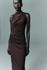 Vestido midi de color negro, con cuello subido y diseño sin mangas. Presenta un detalle de drapeado lateral en la parte delantera y cierre de cremallera oculta en la espalda.