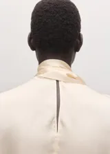 Blusa de seda color crema con cuello alto y detalle de pañuelo integrado en el escote. Presenta un diseño fluido y elegante de manga larga.