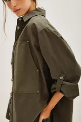Camisa de estilo militar confeccionada en canvas de algodón, con corte oversize, cuello clásico, cierre frontal con botones y bolsillos laterales. Presenta aberturas en los costados y mangas con ajuste de botón.