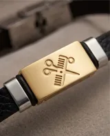 Pulsera de diseño profesional con placa central rectangular de acero dorado que presenta un grabado de tijeras. La estructura combina la placa con segmentos de cuerina negra y detalles metálicos, ofreciendo un estilo moderno y temático.