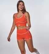 Conjunto deportivo de dos piezas color naranja, compuesto por un top de escote redondo con costuras a la vista en contraste y un short de tiro alto con costuras a la vista. El top presenta el logo de la marca estampado en el frente.