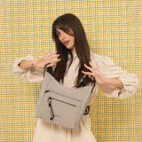 Cartera bandolera gris de formato amplio, con correa larga regulable y desmontable con inscripciones. Cuenta con cierre superior, bolsillo frontal con cierre y detalle colgante de cordones, y un bolsillo trasero con cierre.