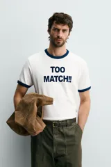 Camiseta de corte regular con cuello redondo y mangas cortas con ribetes en contraste de color. Presenta una estampa frontal con la frase 'TOO MATCH!!' en tipografía destacada.