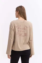 Remera de manga larga color beige con silueta relajada y mangas amplias. Presenta una estampa en la espalda con la frase 'Be kind to your mind' acompañada de ilustraciones de un cupido, flores y lazos en tono bordó.