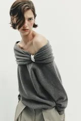Jersey de punto color gris oscuro, con escote recto tipo off-shoulder, manga larga y detalle de aplique metálico en el hombro. Presenta acabados en rib en puños y ruedo.