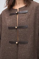 Capa de lana fina sin teñir, con diseño de corte amplio y mangas tipo kimono. Presenta cierre frontal mediante tres alamares de cuero en tono marrón.