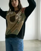 Remera tipo poncho de silueta amplia y relajada, confeccionada en una combinación de jersey y viscosa. Presenta un diseño bicolor con un panel frontal estampado con formas orgánicas en tonos tierra y verde, y mangas y espalda en color negro.