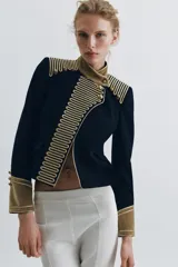 Chaqueta de estilo militar en color negro, con cuello subido y puños en terciopelo color verde oliva. Presenta detalles de alamares dorados en el frente y hombros, bolsillos con solapa y cierre frontal con botones metálicos en relieve.