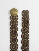 Cinturón de cuero sintético marrón oscuro, compuesto por una tira de círculos grandes adornados con ojales dorados y remaches, y una hebilla circular grande con relieve floral en tono dorado envejecido.