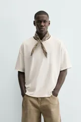 Camiseta de corte relajado (relaxed fit) color blanco roto, con cuello redondo y manga corta. Presenta un bordado sutil en el pecho con diseño floral en tonos claros.