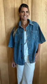 Sobrecamisa de denim soft en tono azul medio, con corte oversize, mangas cortas, cuello clásico y cierre frontal con botones metálicos. Presenta dos bolsillos frontales con solapa y un ruedo redondeado.