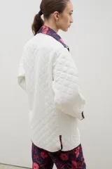 Campera acolchada de corte holgado, color blanco con diseño capitoneado en forma de rombos. Presenta cuello camisero y bordes internos con estampado floral en tonos azul y rojo, cierre frontal con botones a presión y bolsillos laterales.