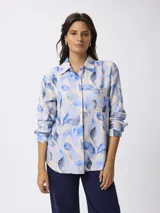 Camisa de manga larga con cuello clásico y cierre frontal de botones. Presenta un estampado de hojas en tonos azules sobre fondo claro y un bolsillo frontal en el pecho.