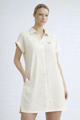 Vestido tipo camisa corto de color beige, confeccionado en viscosa suave y liviana. Presenta cuello camisero, cierre frontal con botones, mangas cortas con dobladillo y bolsillos laterales. Incluye un pequeño bordado de corazón en el pecho.