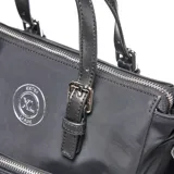 Cartera tipo tote de tela técnica color negro, con doble asa de mano y correa de hombro ajustable. Presenta múltiples bolsillos frontales con cierre metálico, logo circular de la marca en el frente y detalle de cordón decorativo con argollas.