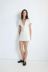 Vestido corto de Zara, color blanco, con escote en pico, mangas cortas abullonadas y detalle de lazo frontal en el mismo tejido. Presenta un corte entallado y cierre de cremallera oculta en la espalda.