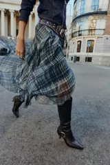 Bota de caña alta en color marrón oscuro, confeccionada en cuero con acabado liso. Presenta un diseño de punta fina, tacón alto fino y una tira decorativa en el tobillo con una hebilla metálica dorada. Incluye cierre lateral mediante cremallera.