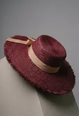 Sombrero de paja tejido en color bordó, con ala ancha y borde deshilachado. Presenta una cinta de tela color beige atada en un moño alrededor de la copa.