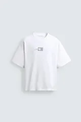 Camiseta de corte boxy fit, color blanco, con cuello redondo y manga corta. Presenta una aplicación de etiquetas decorativas a contraste en el centro del pecho.