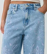 Pantalón wide leg de jean celeste con talle medio y bordados florales en hilo celeste claro a lo largo de las piernas. Cuenta con bolsillos funcionales, pasadores para cinto y cierre frontal con botón y cremallera.