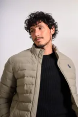 Campera puffer de corte clásico en color beige, con cierre frontal, cuello alto y puños elásticos. Presenta un diseño acolchado horizontal y un pequeño logo bordado en el pecho.