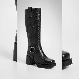 Bota de caña alta confeccionada en cuero vacuno, con diseño de punta cuadrada y plataforma. Presenta un taco cuadrado de madera, cierre lateral interno y una tira decorativa con herraje metálico circular en el tobillo.