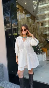 Vestido corto de broderie color blanco, con cuello camisero, cierre frontal de botones y mangas tres cuartos. Presenta un diseño de corte holgado con falda de volados y ruedo irregular.