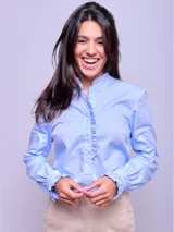 Camisa de algodón color celeste, con cuello alto, cierre frontal de botones y detalles de volados verticales a lo largo de la botonera. Presenta mangas largas con puños ajustados y terminación con volados.