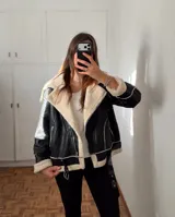 Campera tipo gamulán de cuero sintético negro con interior y detalles de corderito sintético en color crema. Presenta cierre frontal, bolsillos laterales con cierre y cinturón ajustable en el ruedo.