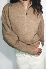 Jersey de punto melange color beige, con cuello subido, cierre frontal con cremallera y manga larga con hombros caídos.