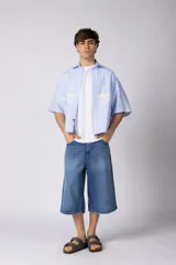 Camisa de manga corta de corte boxy, confeccionada en popelina a rayas celestes y blancas. Presenta cuello camisero clásico, cierre frontal con botones y estampado tipográfico en el pecho.