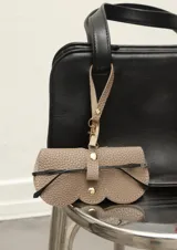 Funda para lentes color beige con textura símil cuero, cierre con broche a presión y correa con mosquetón para colgar de la cartera.