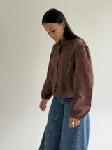 Blusa de brodery color blanco, con cuello redondo, mangas largas abullonadas con puños elásticos y cierre frontal con botones forrados. Presenta detalles de puntillas verticales en el frente.