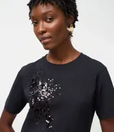 Remera de algodón de corte recto, con cuello redondo y mangas cortas. Presenta un diseño decorativo de lentejuelas bordadas en la parte frontal.