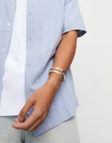 Pulsera rígida de aleación de metales bañada en plata de ley, con diseño abierto y logo de la marca grabado en uno de los extremos.