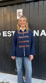 Campera de estilo militar en color azul marino, confeccionada en tejido de punto grueso. Presenta un diseño de cuello redondo, cierre frontal con tiras de cuero marrón y botones metálicos decorativos. Posee un corte recto y holgado.