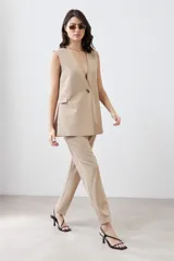 Chaleco largo de corte sastrero en color beige, con escote en V profundo, cierre frontal con un botón metálico y bolsillos laterales con solapa.